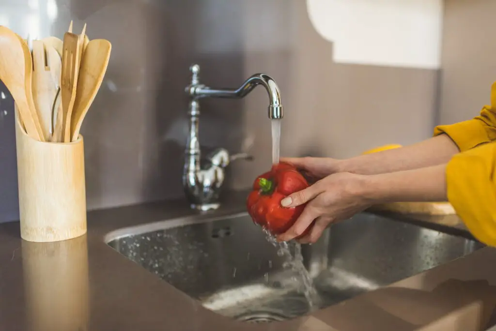 An image of a Woman Washing Red Bell Pepper in the Kitchen Sink.