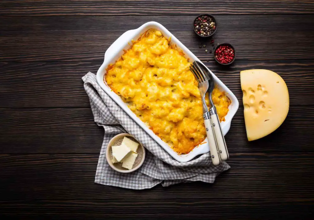An image of baked mac and cheese in white casserole on rustic wooden background, with seasonings, butter, spoon and fork.