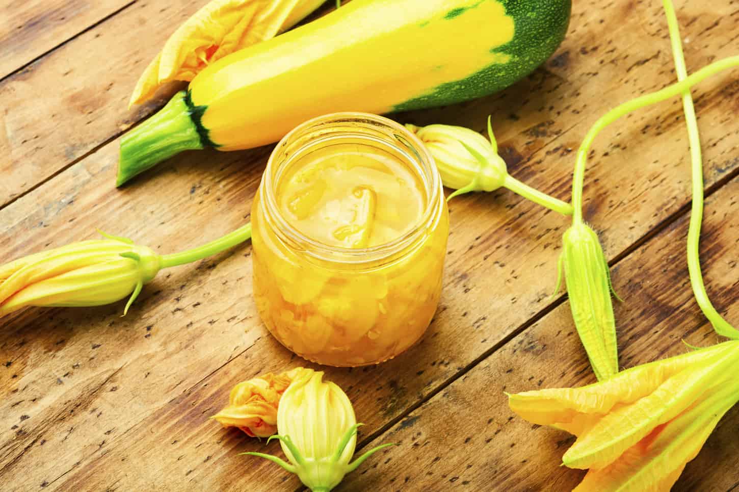 An image of a bottle of zucchini jam, a zucchini turning yellow, and zucchini flowers on a wooden table.