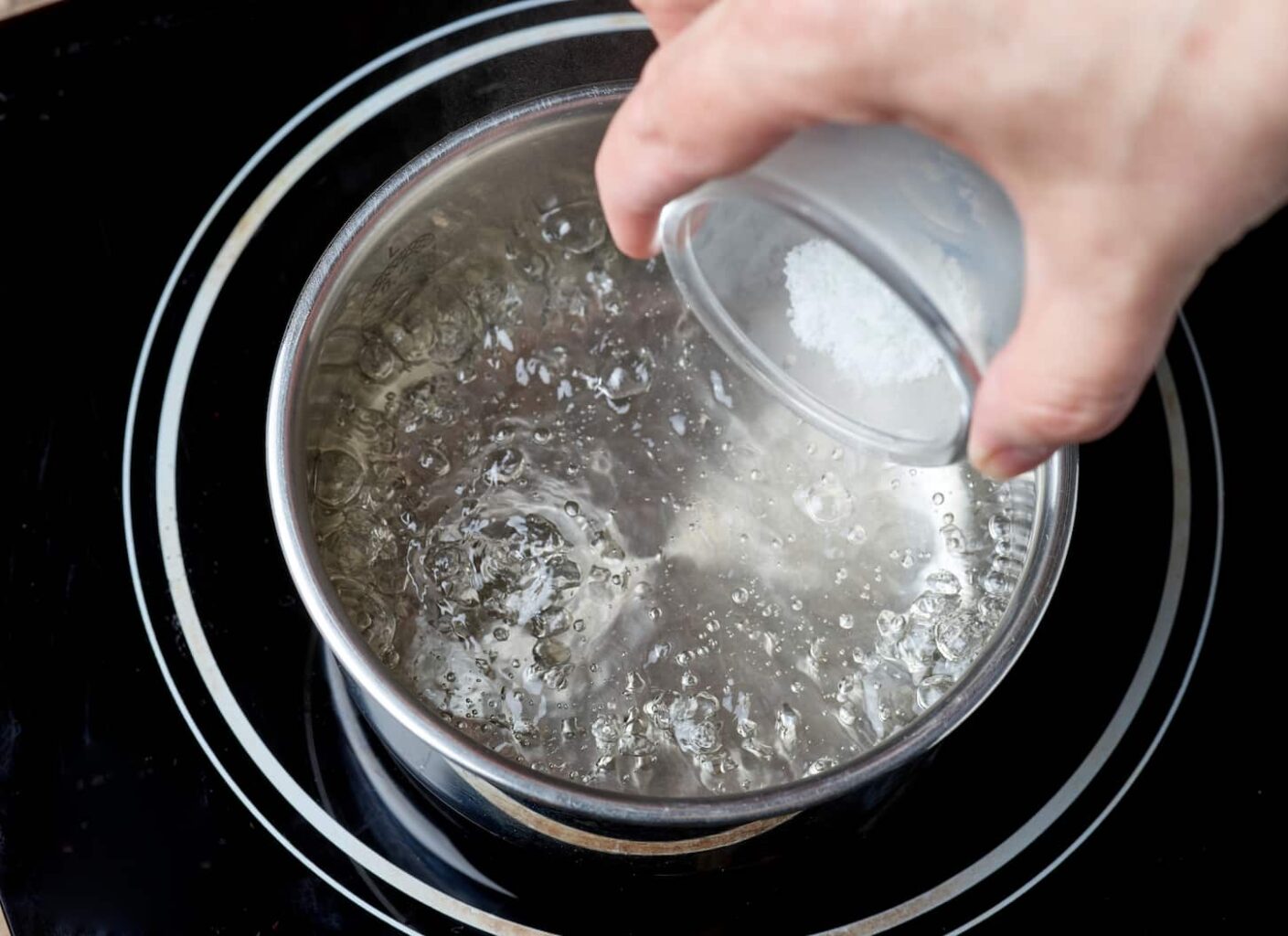 An image of a hand adding salt to a boiling water pot on an electric induction hob.