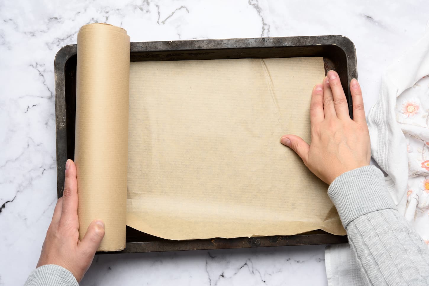 An image of two female hands covered with brown parchment paper on an iron rectangular baking sheet.