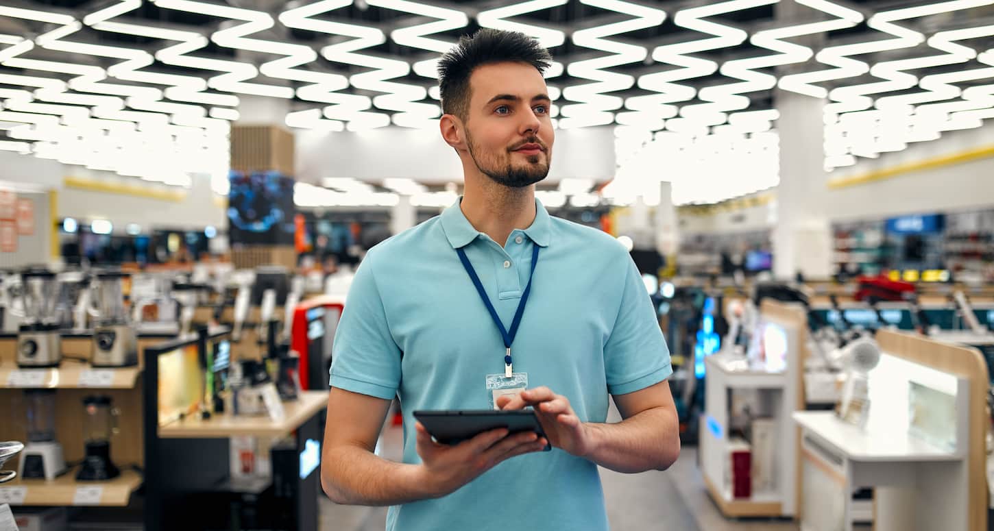 An image of a young successful sales assistant in uniform with a tablet in his hands against the background of the interior of the hall in a home appliances and electronics store.