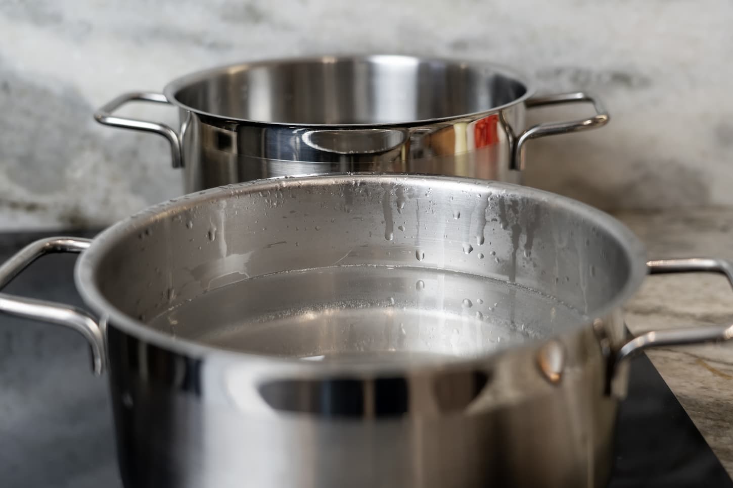 An image of Stainless steel pots filled with water on a black ceramic hob and marble countertop.