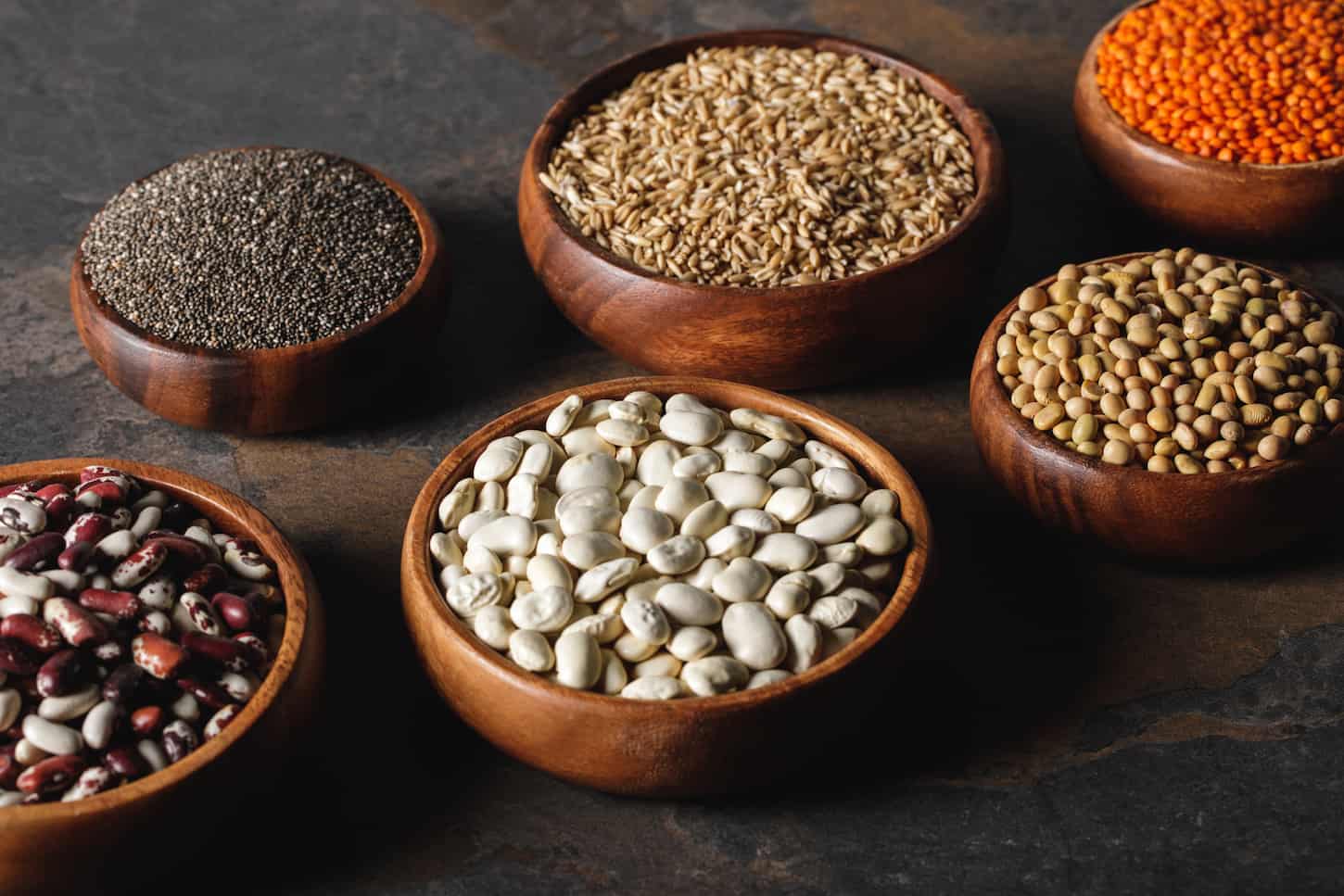 An image of various beans, chia seeds, and oat groats in wooden bowls on the table.