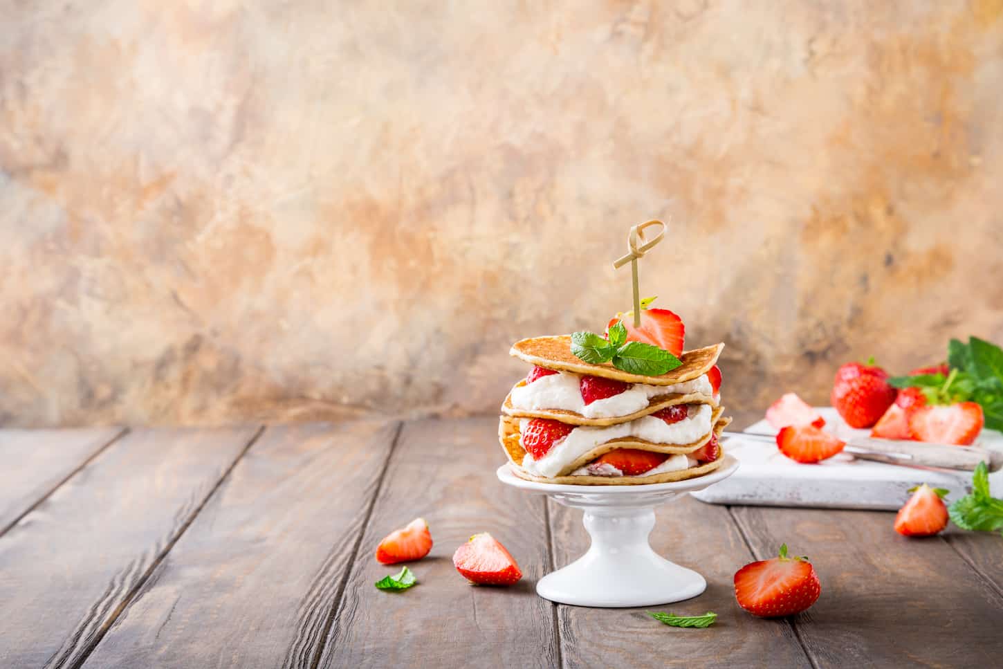 An image of a Small cake made of pancakes with cream and strawberries on a white porcelain cake stand.