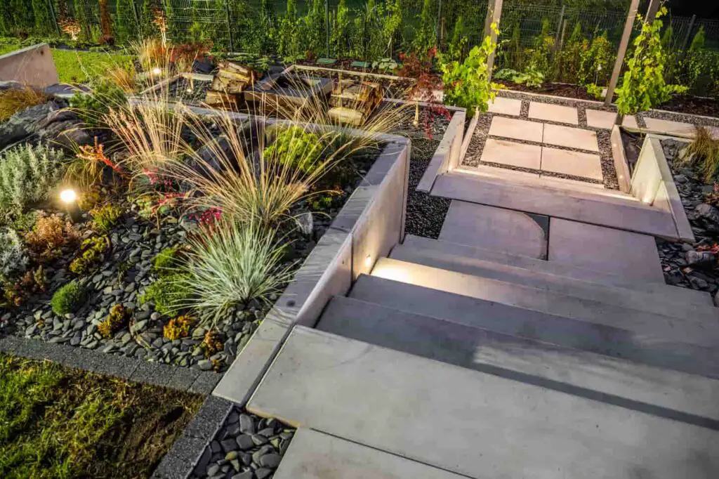 An image of a Professionally Landscaped Backyard Garden with Concrete Stairs and Large Tiered Flowerbeds Decorated with Pebbles and Outdoor Lighting Lamps.