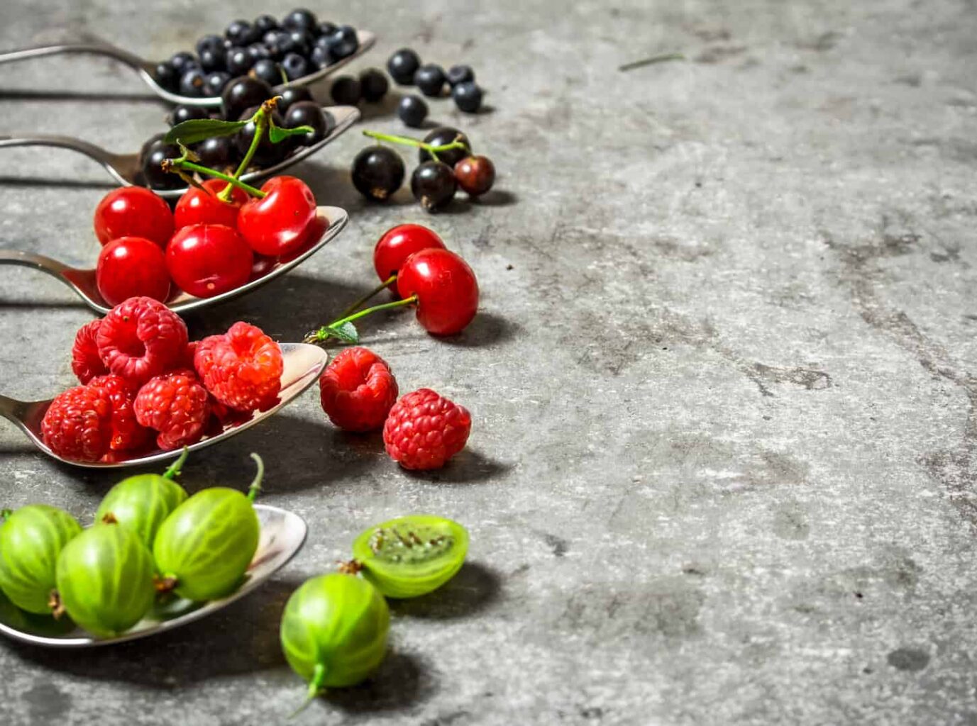 An image of different berries placed in spoons on a granite countertop.