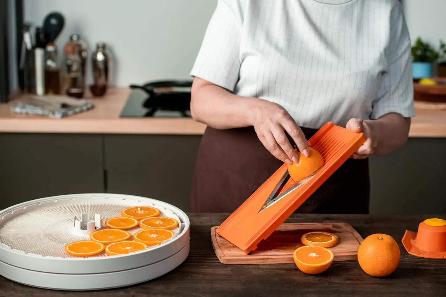 An image of an unrecognizable woman standing at home kitchen and cutting oranges on slicer while dehydrating it at home.