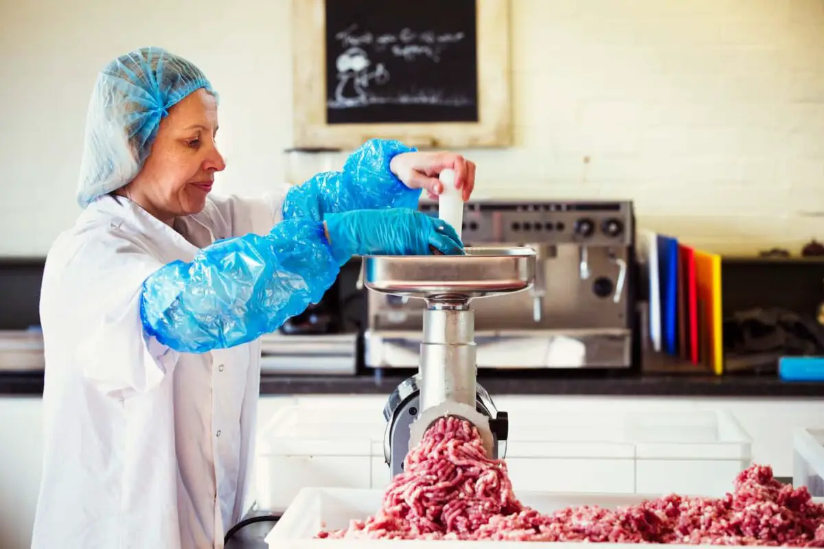 An image of a Woman working in a butchery, wearing protective clothes and gloves, putting minced meat into a meat grinder.